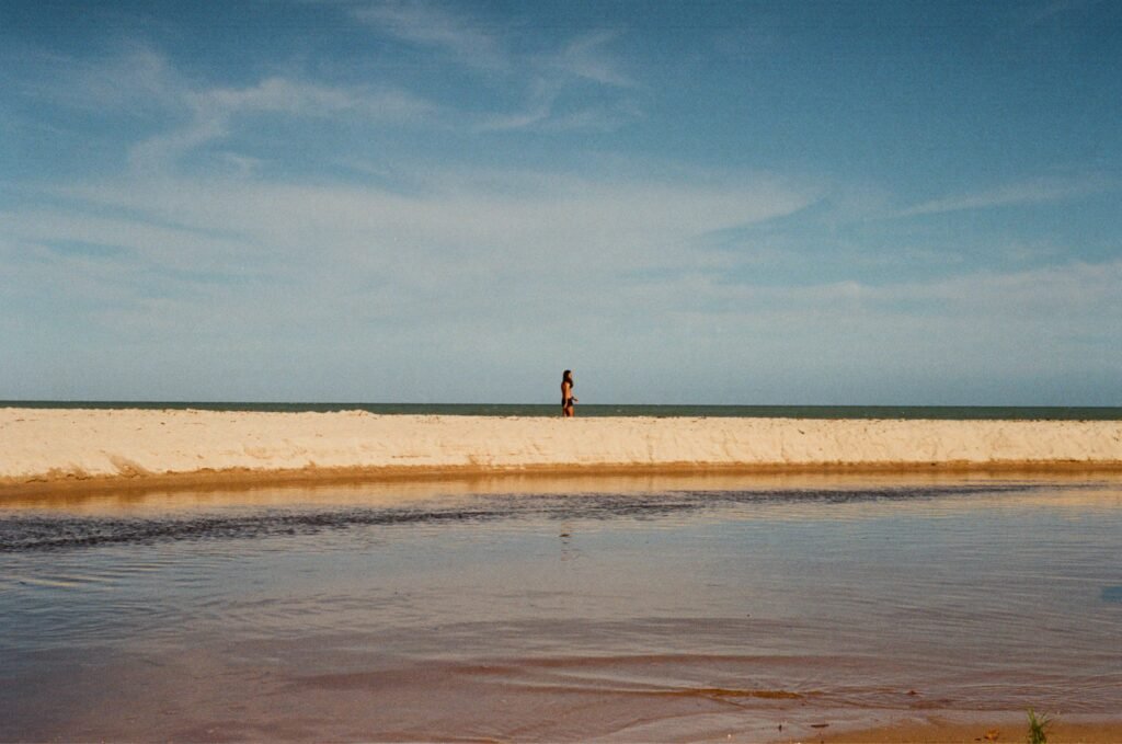 An adult walking on a tranquil sandy beach under a blue sky in Brazil.